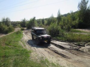 Off-road vehicle driving on a sandy dirt road through a green, wooded area with power lines overhead.