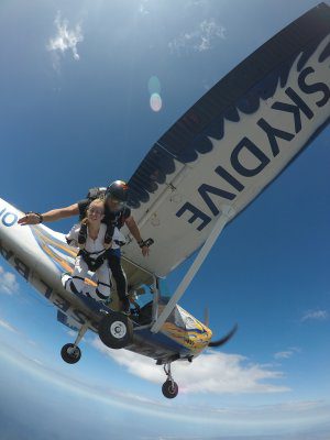 Two tandem skydivers jump from a small plane into clear blue sky, airplane wing reading'SKYDIVE' in the background.