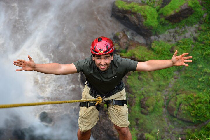 Man in a red helmet and harness suspended by a rope, arms wide, above a waterfall and green cliffside.