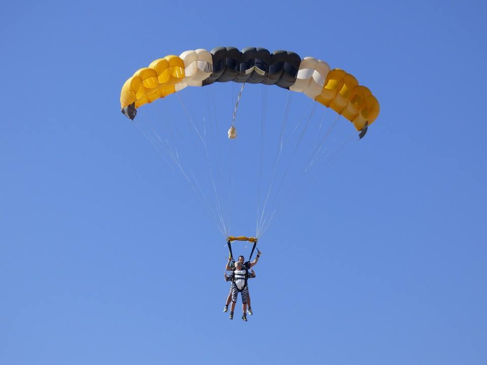 Two people tandem-skydiving under a multi-color parachute (yellow, black, white) against a clear blue sky.