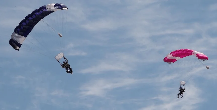 Two people paragliding under open parachutes in a blue sky, one with a blue-purple canopy on the left and a pink canopy on the right.