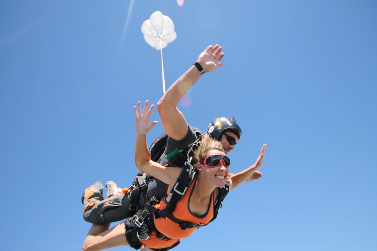 Tandem skydiving pair in free fall: passenger in an orange top with red goggles, instructor behind, both in harness against a blue sky.
