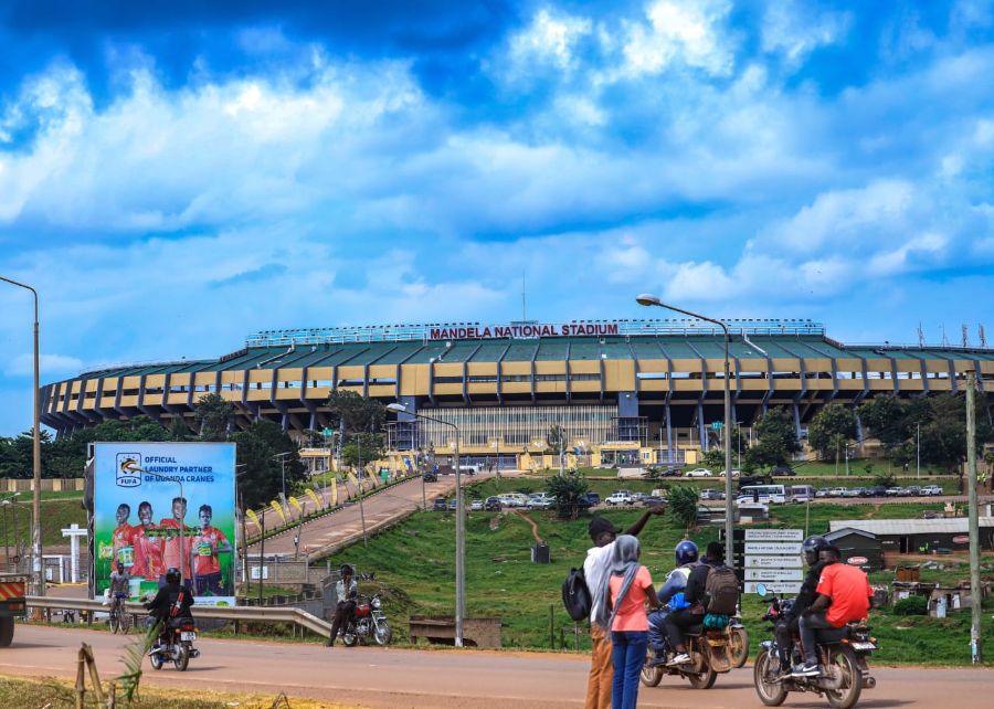 Wide view of Mandela National Stadium with a busy street scene in the foreground—motorcycles, pedestrians, and a billboard under a blue sky.