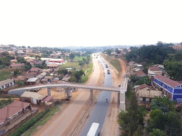 entebbe-mityana | Uganda Car Rental Services Aerial view of a road with a pedestrian bridge crossing a two-lane street, flanked by houses and trees in a small town.