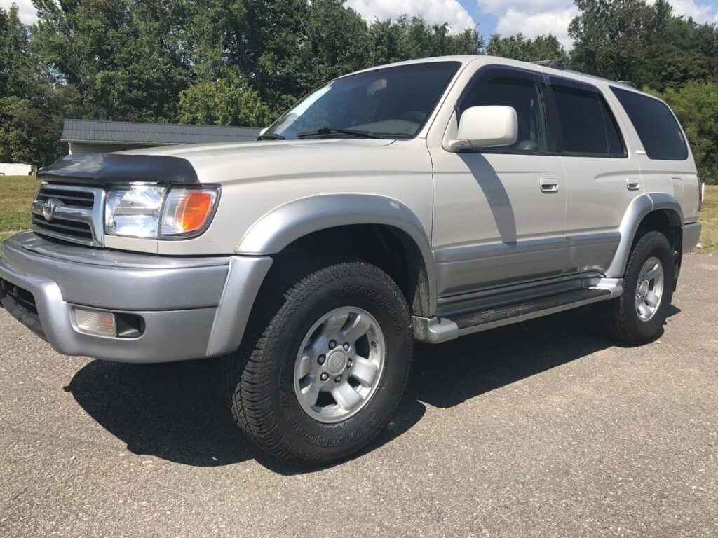 Silver full-size SUV shown from the front-left three-quarter view, parked on a paved lot with trees in the background, featuring large tires and running boards.